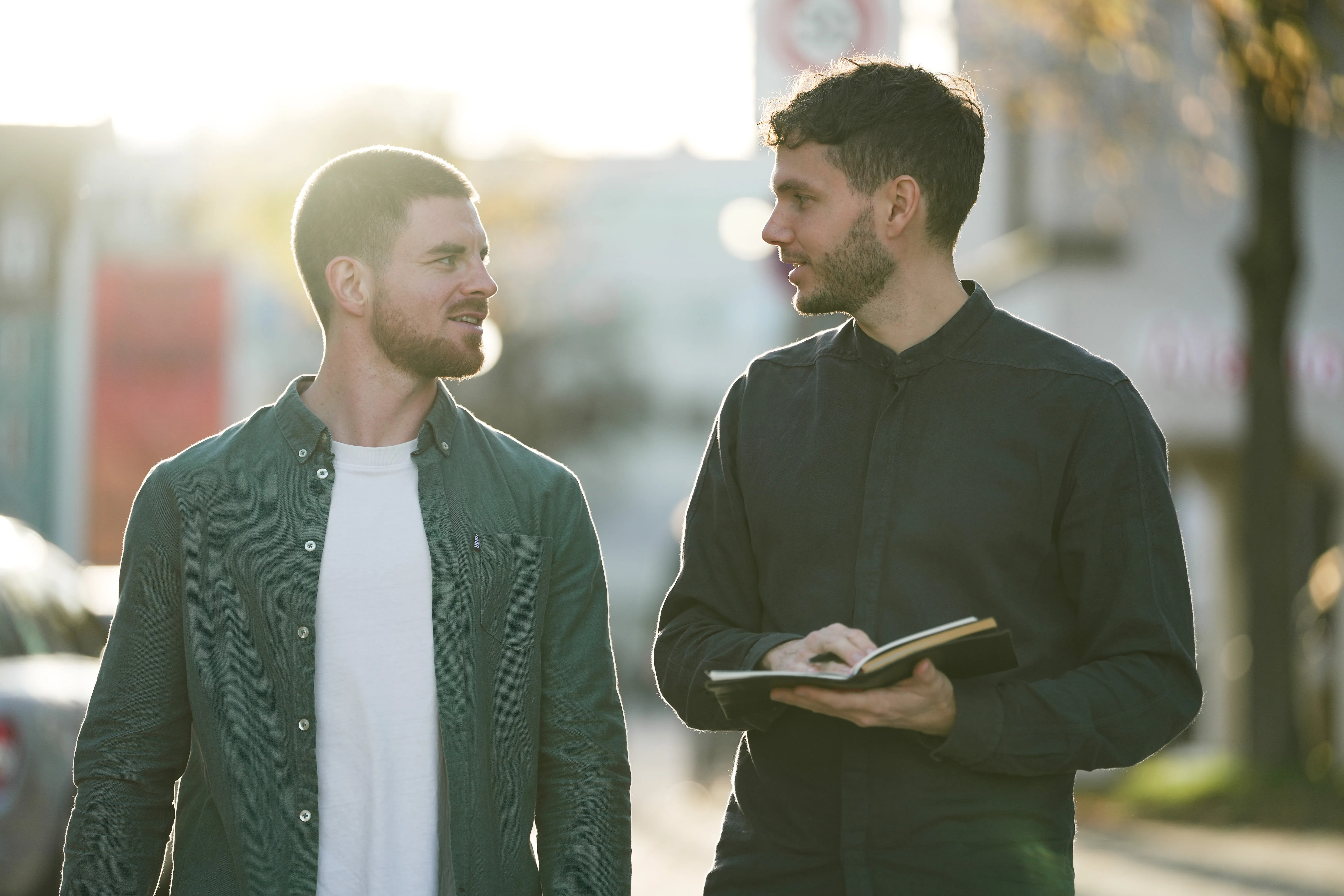 Two men walking outdoors in sunlight, one holding an open notebook and both engaged in conversation.