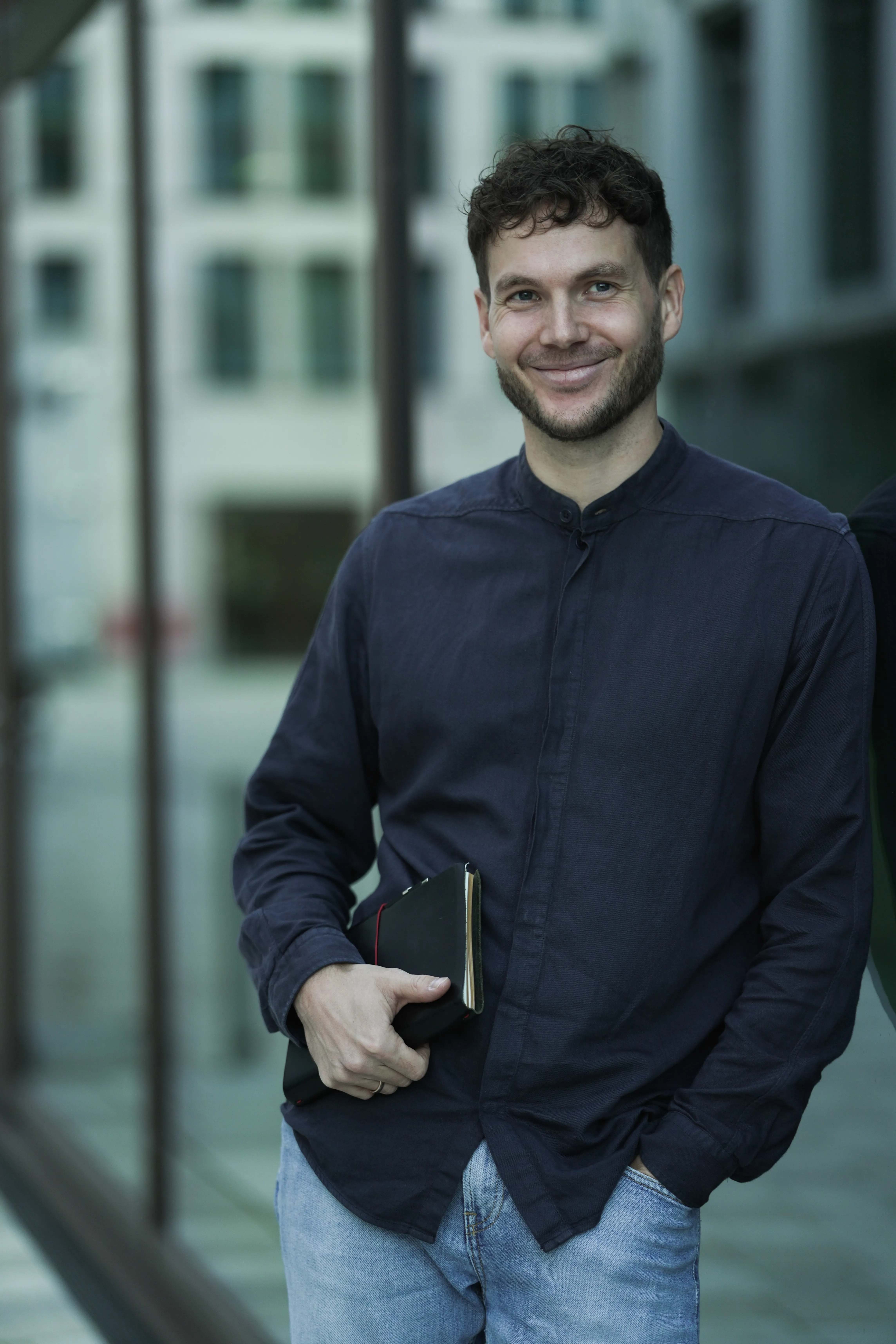 Smiling man with short curly hair wearing a dark buttoned shirt and light jeans holding a black notebook, standing outdoors by glass windows.