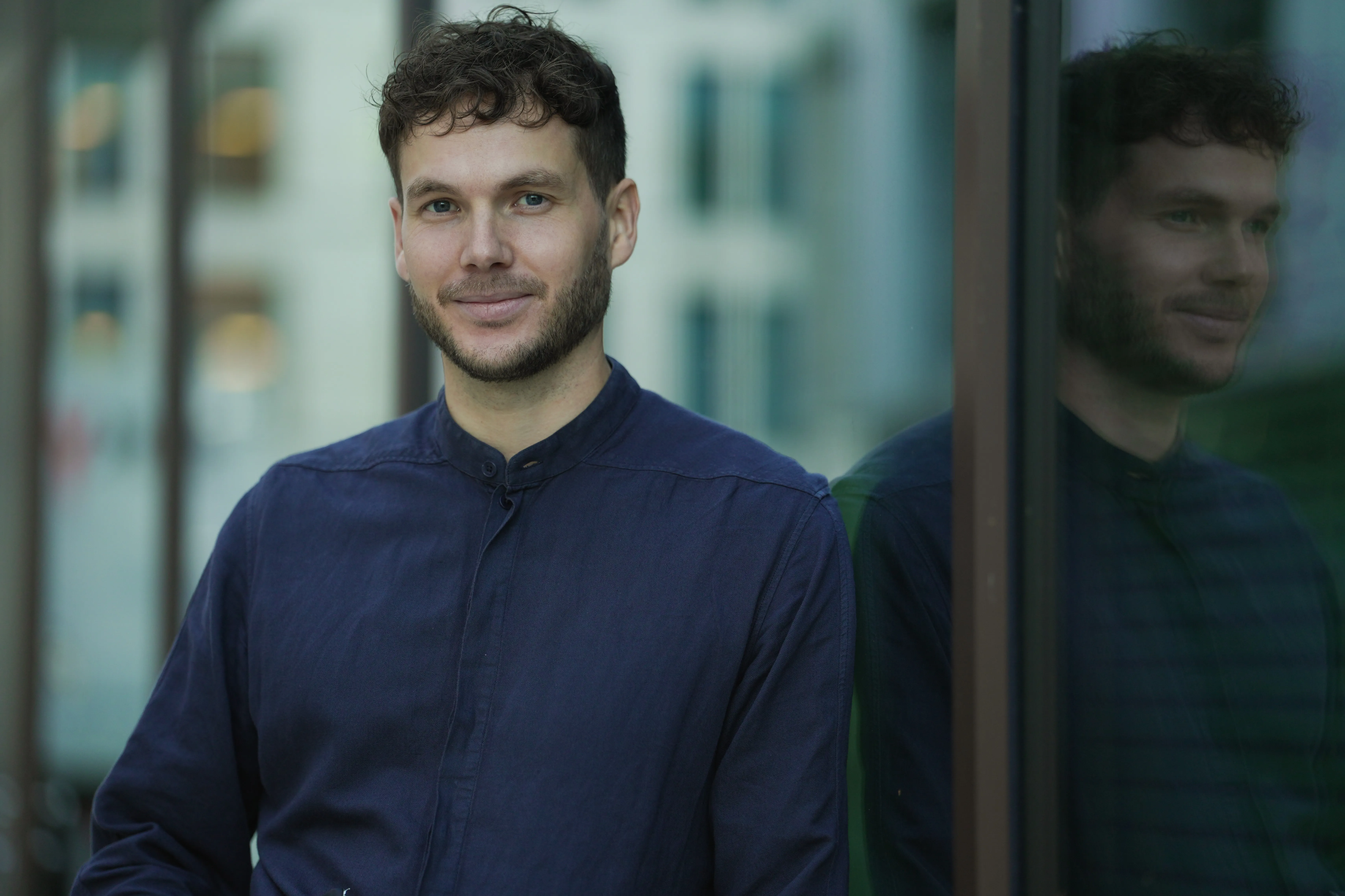 Smiling man with curly hair and beard wearing a dark blue shirt, standing next to a reflective glass wall.